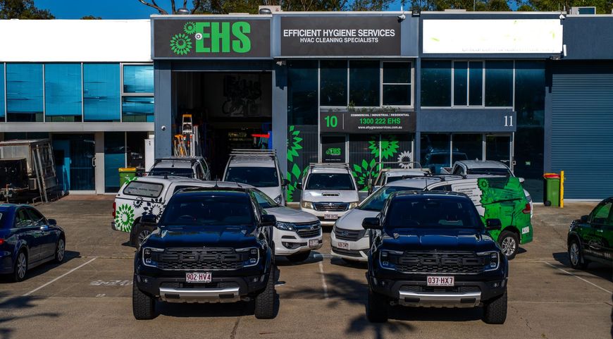 Several vehicles parked in front of a building with EHS sign. Two black trucks in front, vans and cars behind.