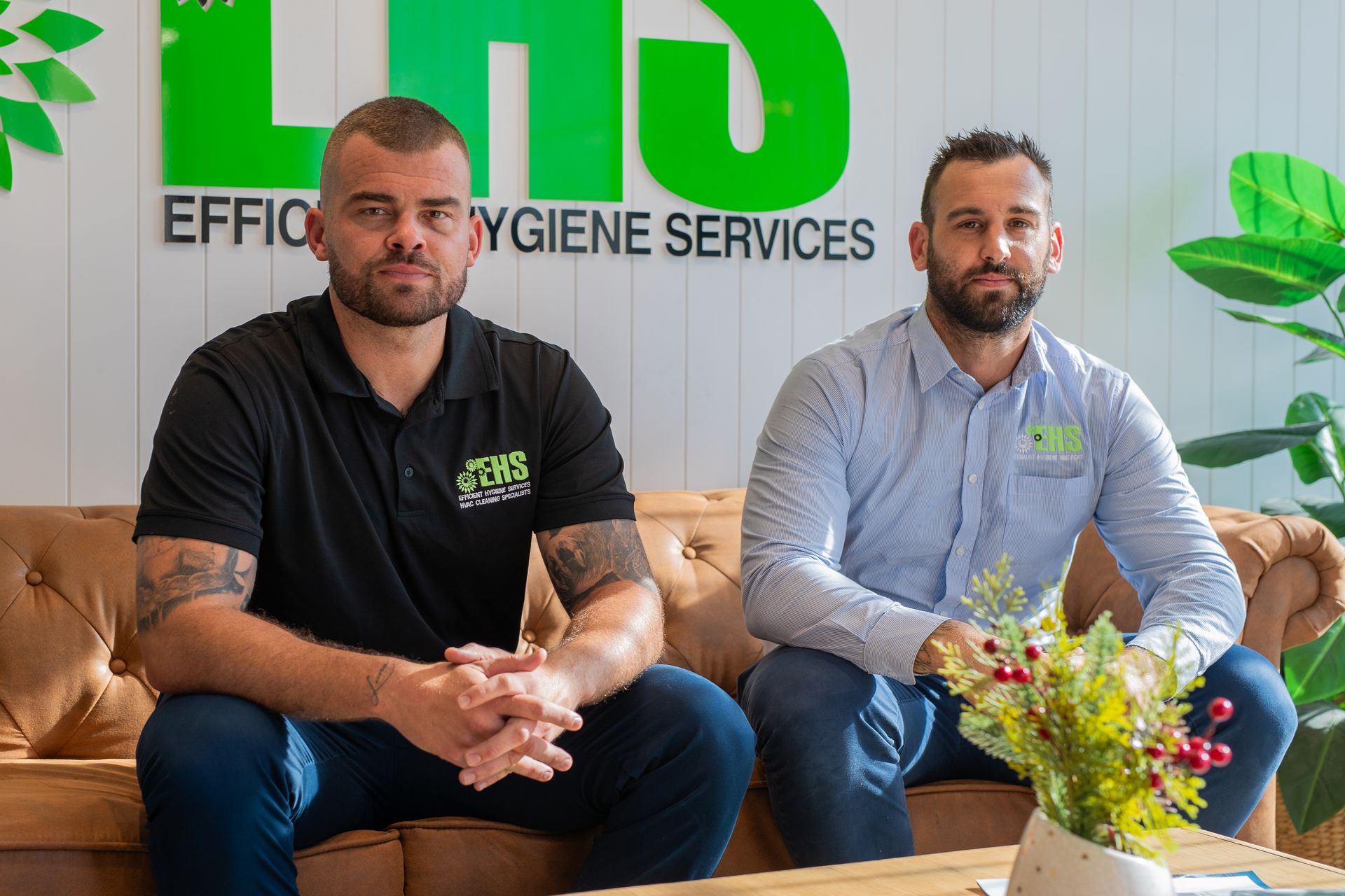 Two men seated on a brown sofa in front of a green and white business sign.