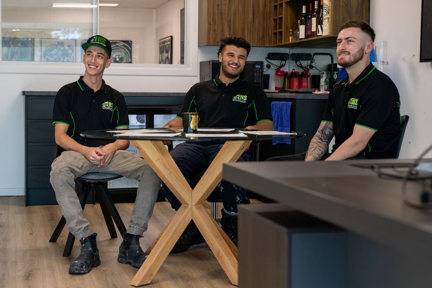 Three people in matching black and green shirts sit at a table in a modern office, smiling.