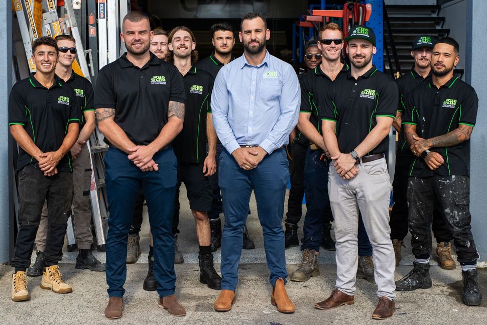 Group of construction workers in front of a building; some wearing black shirts, one in a blue shirt, smiling.