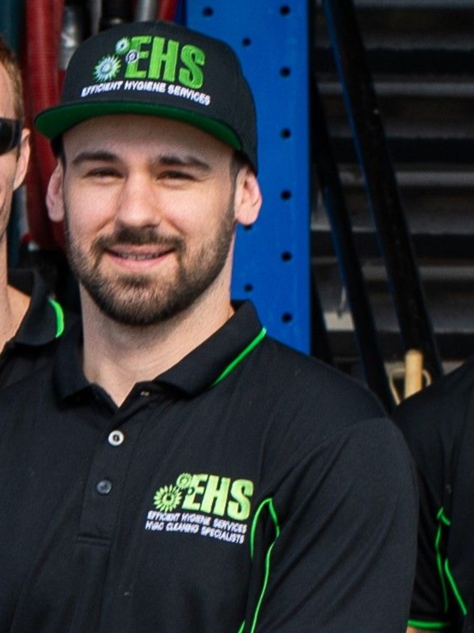 Man wearing a black hat and shirt with a green logo, smiling. Standing outside near tools.