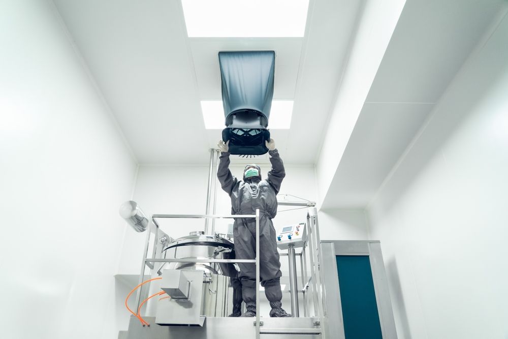 Person in a cleanroom suit working on machinery, reaching up towards a ventilation system.