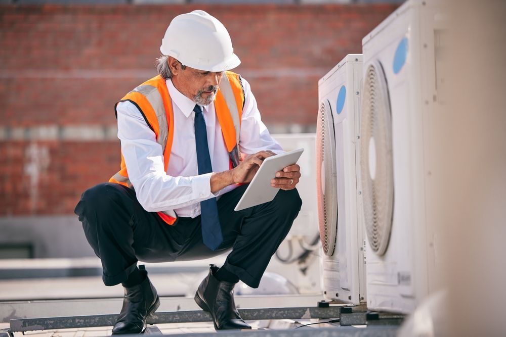 Man in safety gear inspecting HVAC unit on a rooftop, using a tablet.