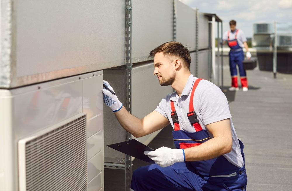 Two workers inspecting HVAC units on a rooftop. One kneels, examining equipment, the other stands in the background.