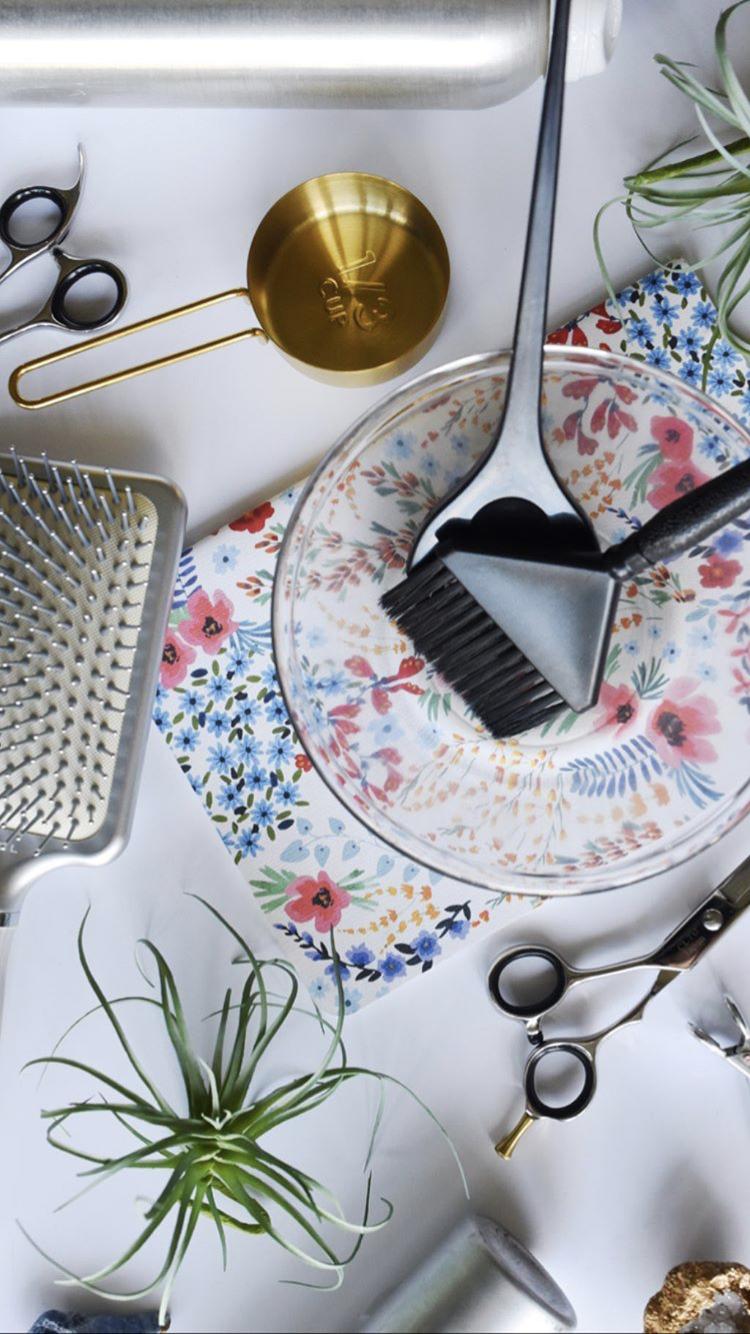 Grooming tools on a floral plate, with scissors, combs, a gold scoop, and small potted plants on a white surface