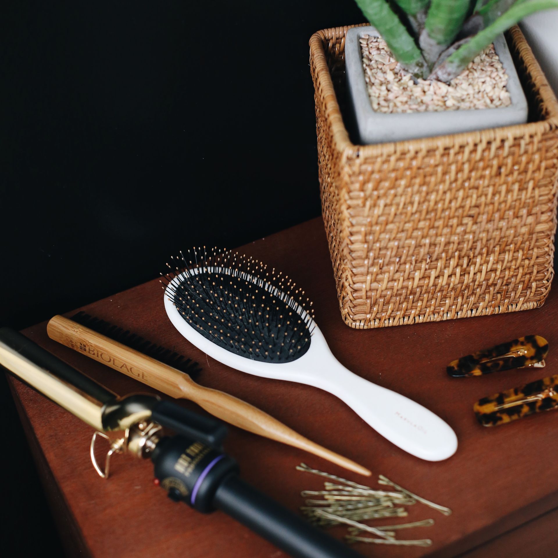 Hairbrush, curling iron, and styling tools on a brown leather surface beside a woven plant pot.