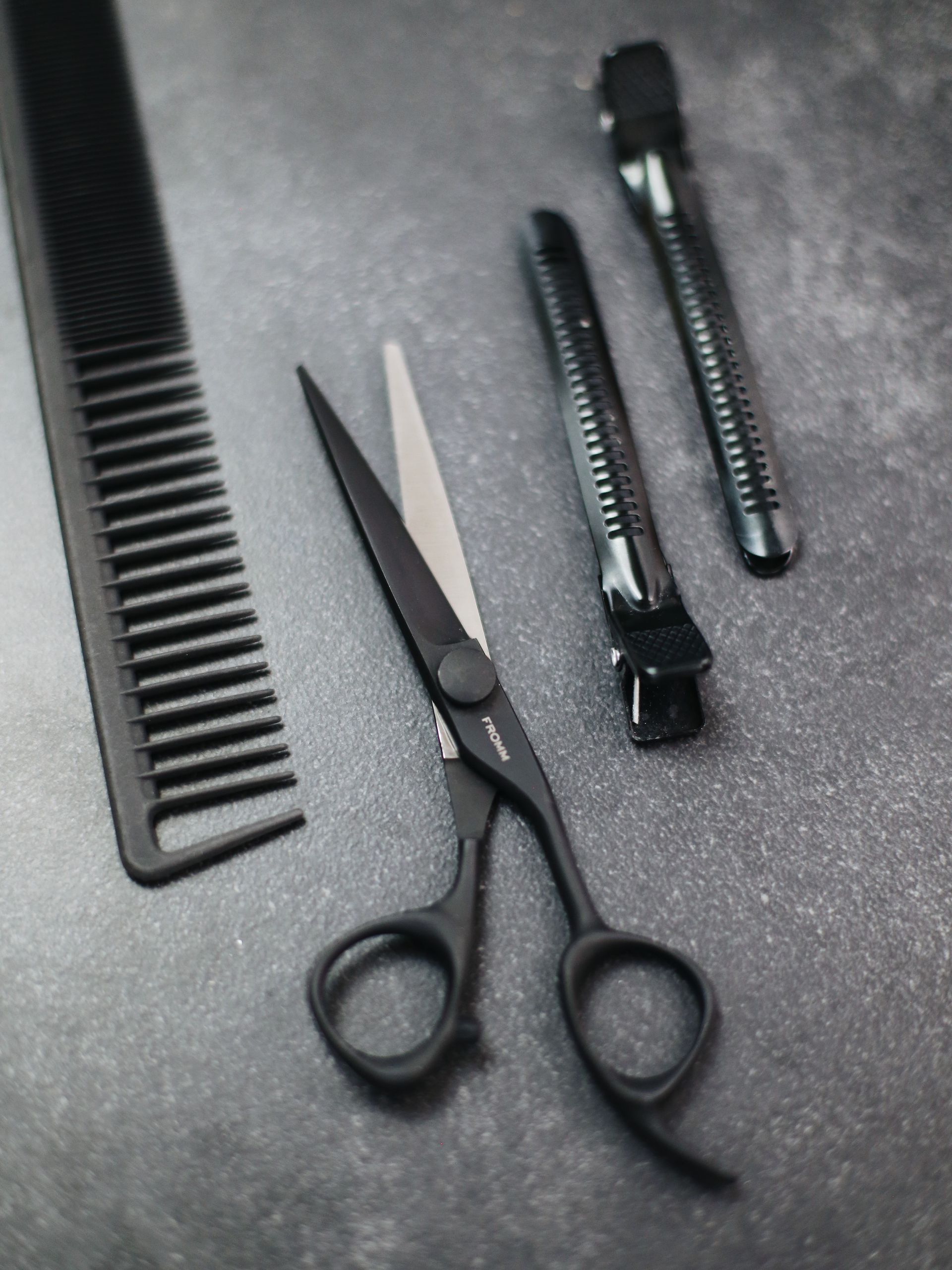Black barber tools: comb, scissors, and two hair clippers on a dark textured surface.