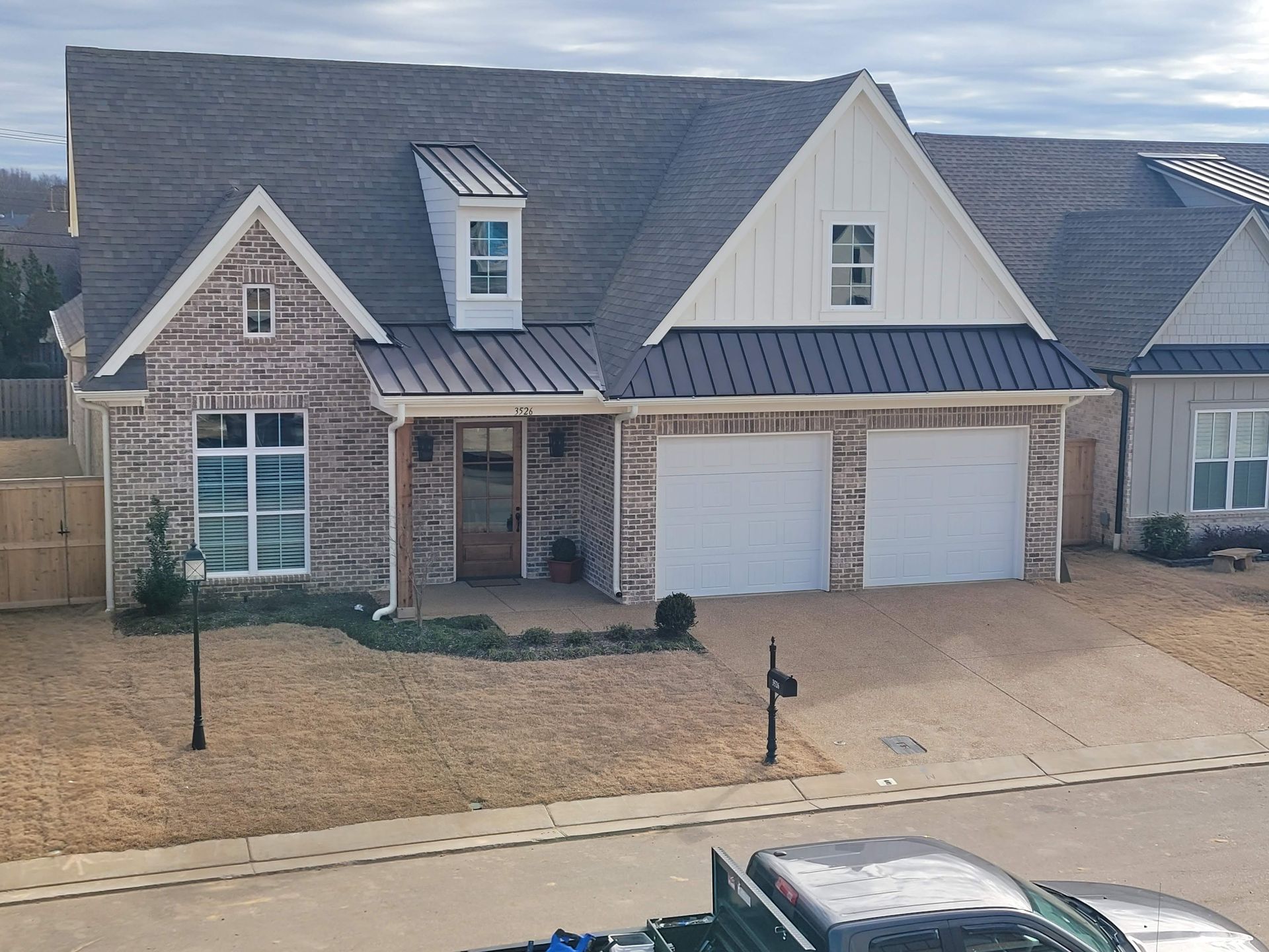 House with brick facade, white gables, and two-car garage. Brown yard, cloudy sky.