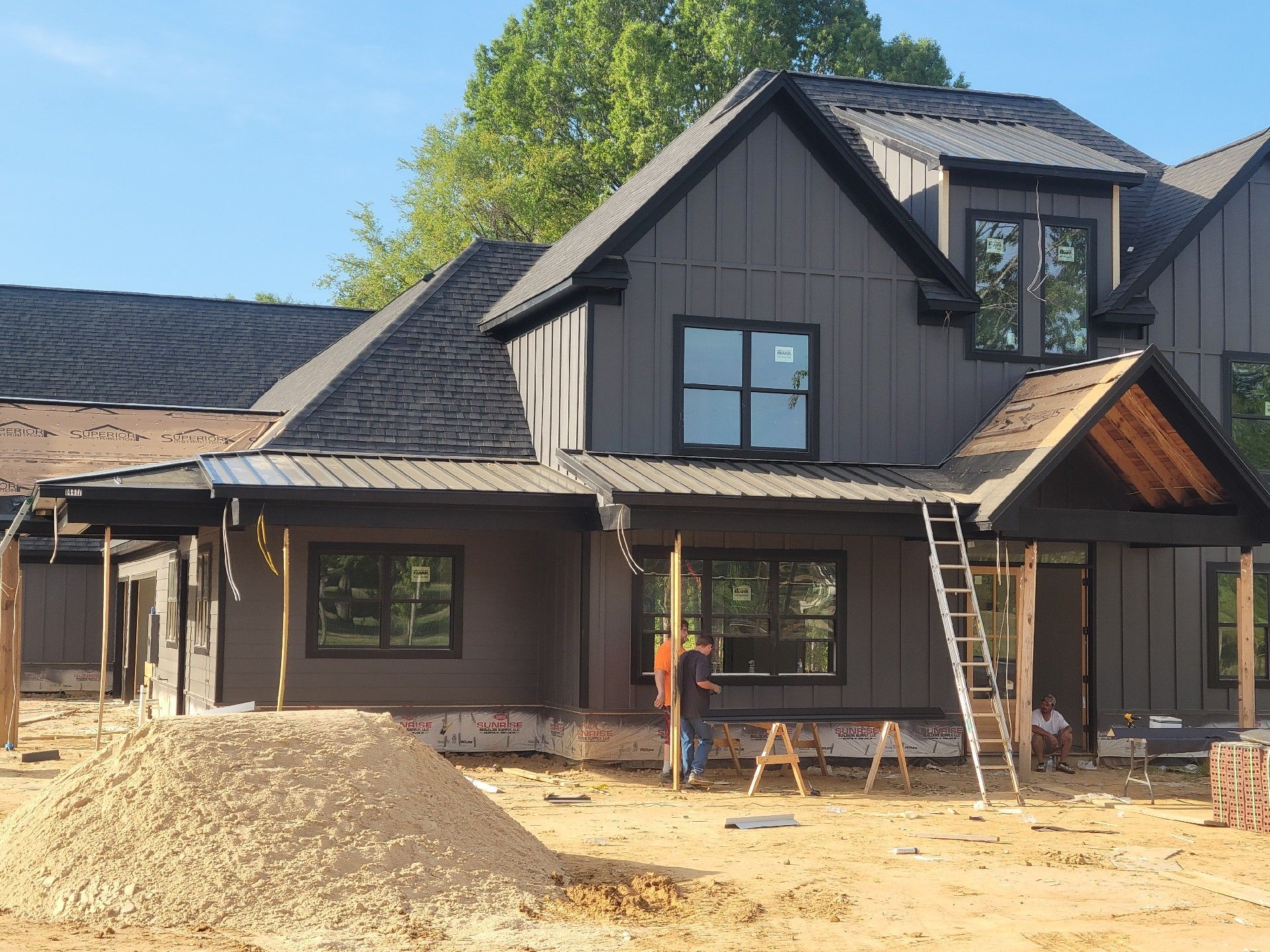Two-story house under construction; gray siding, black roof and trim. Workers on site; dirt pile in foreground.