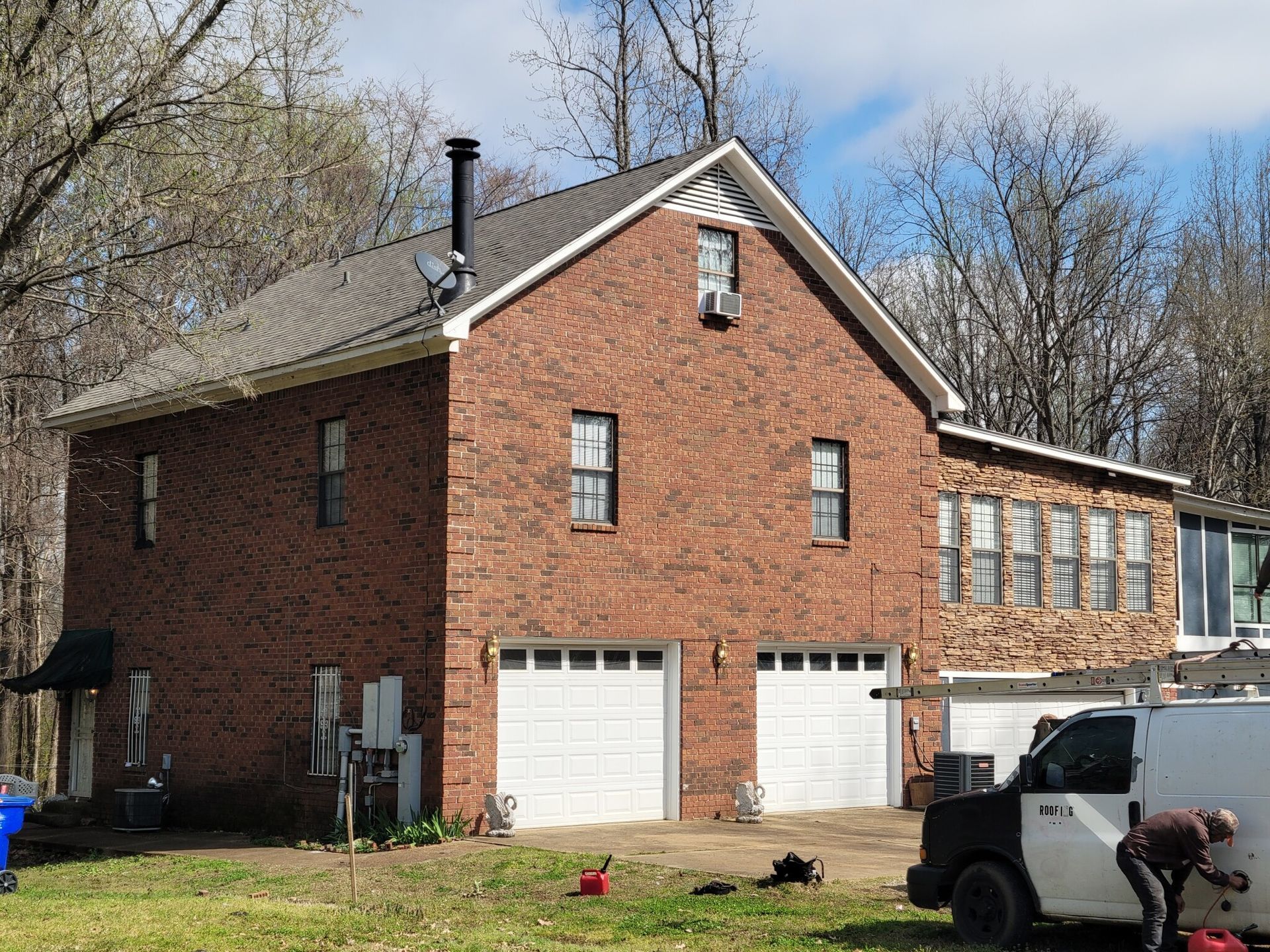 Brick house with two garage doors, windows, and a black roof. A van and a person are in front.