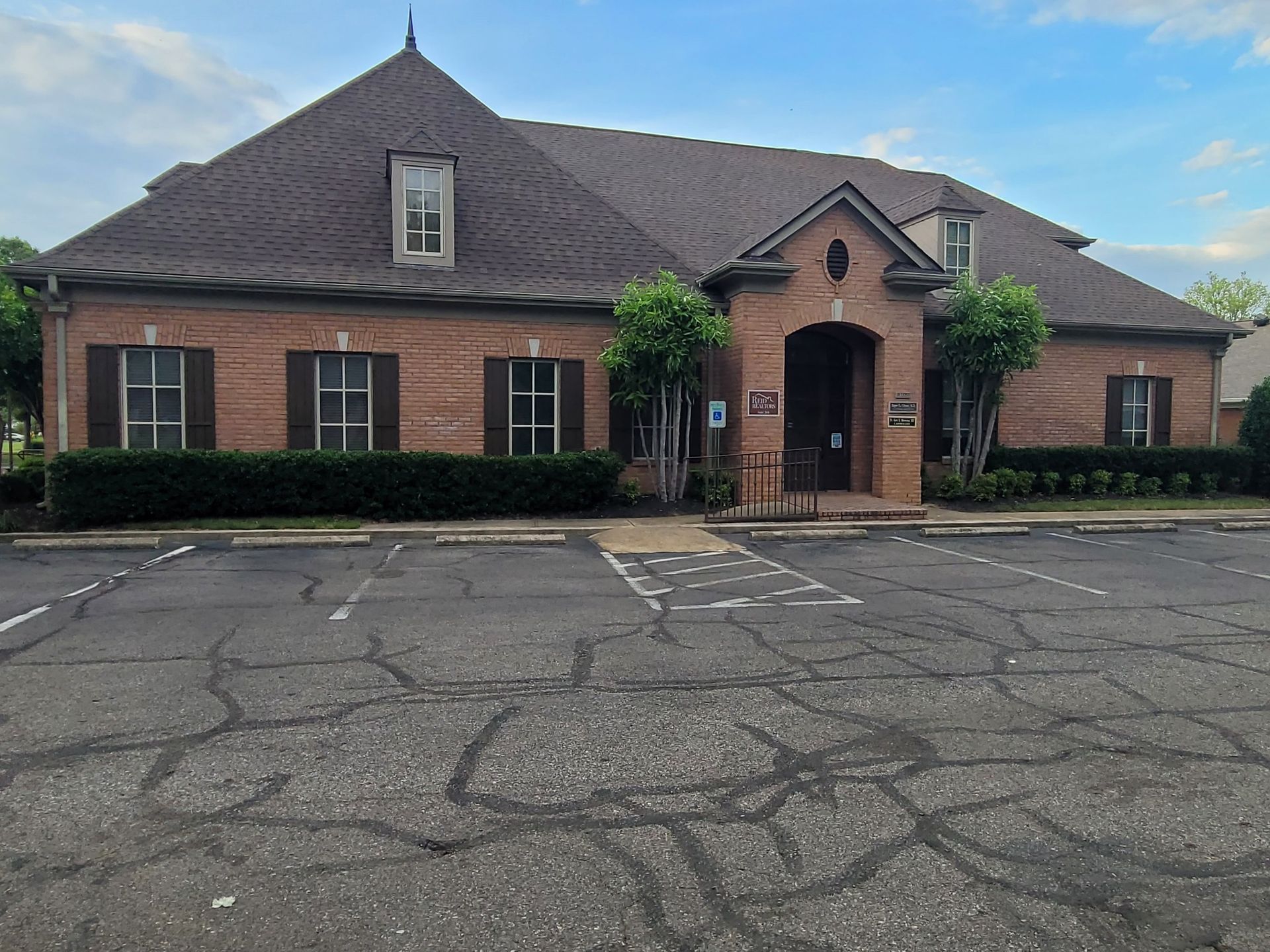 Red brick building with brown roof, windows, and parking lot. Green bushes and trees frame the entrance.