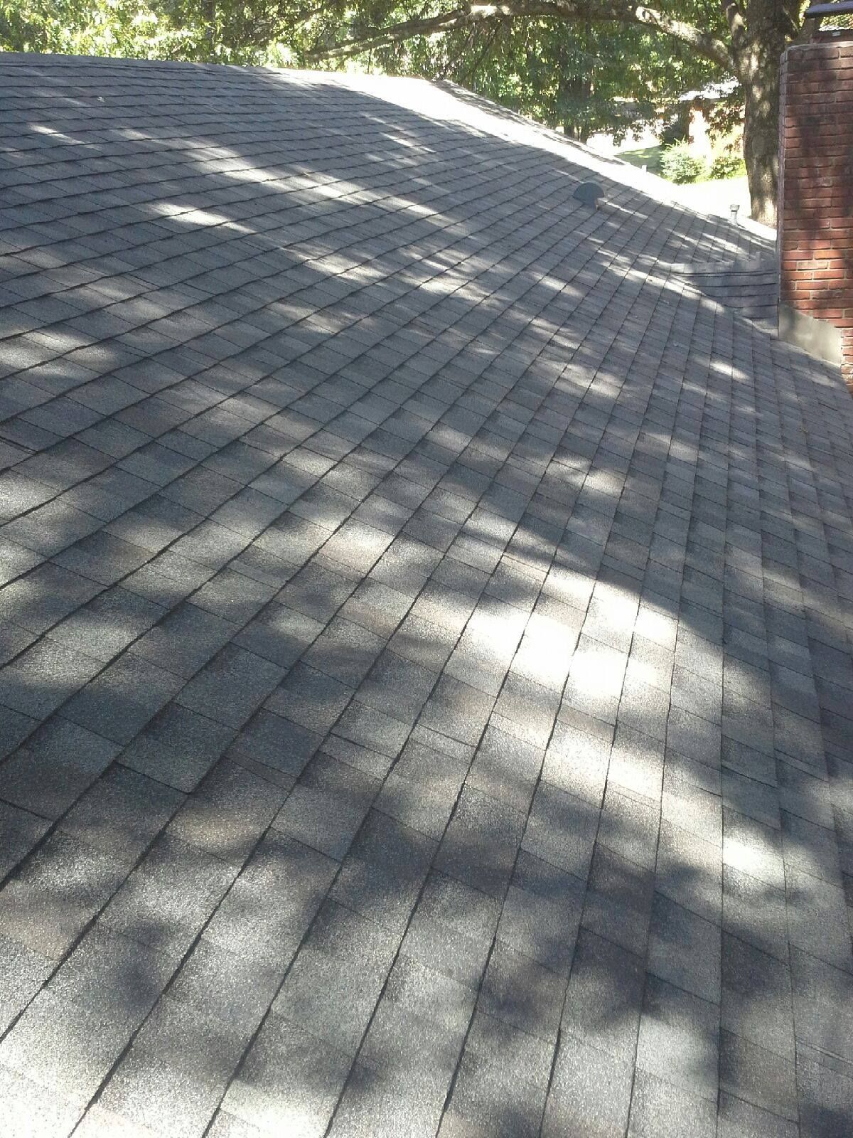 Asphalt shingle roof with dappled sunlight, near a brick chimney and trees.