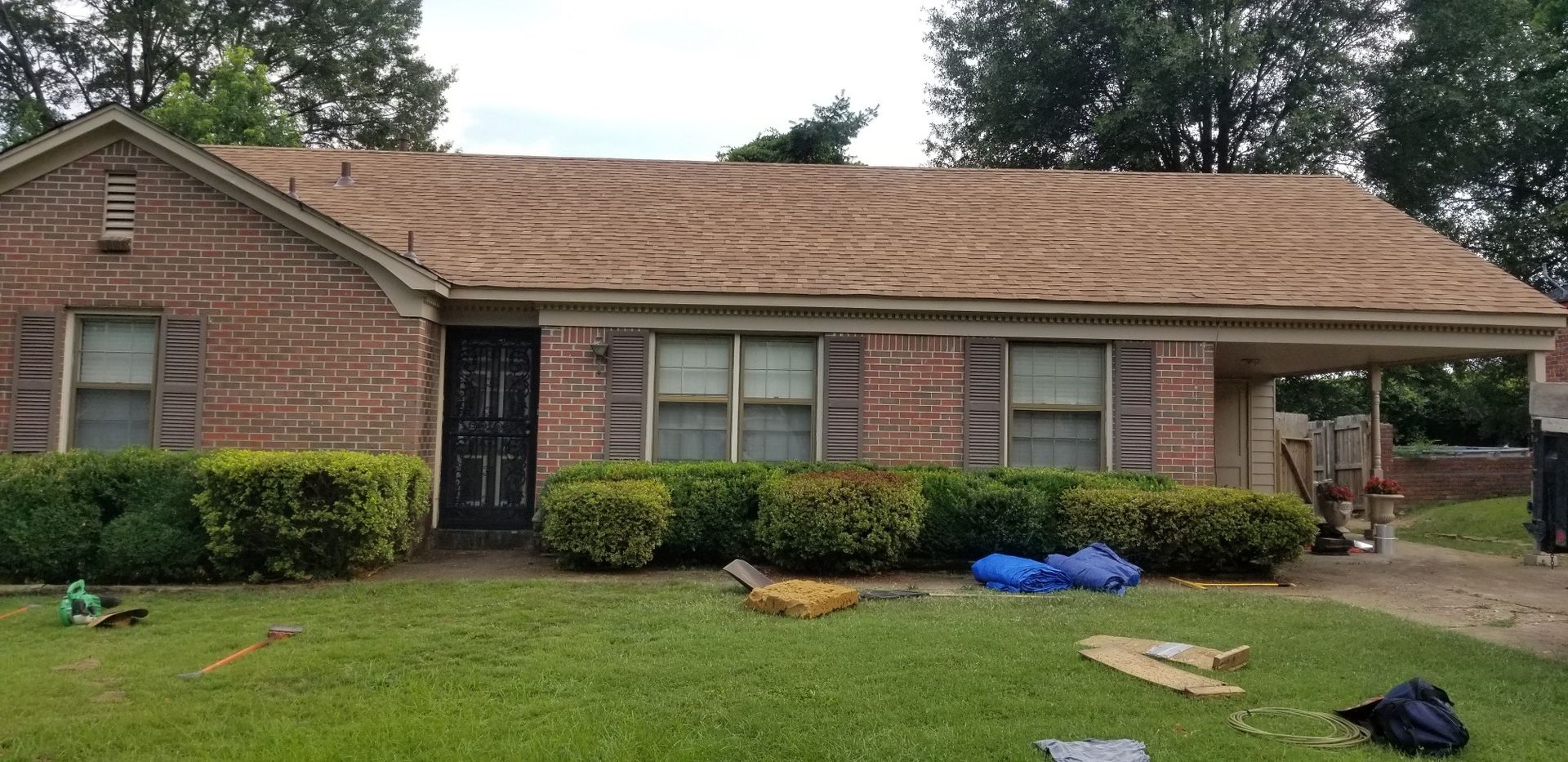 A brick house with a damaged roof, green bushes, and a carport.
