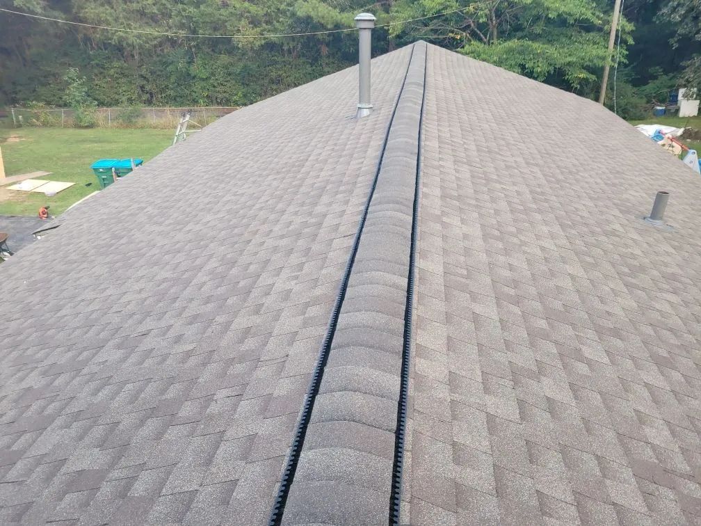 Gray shingled roof with a central ridge vent, viewed against green hillsides