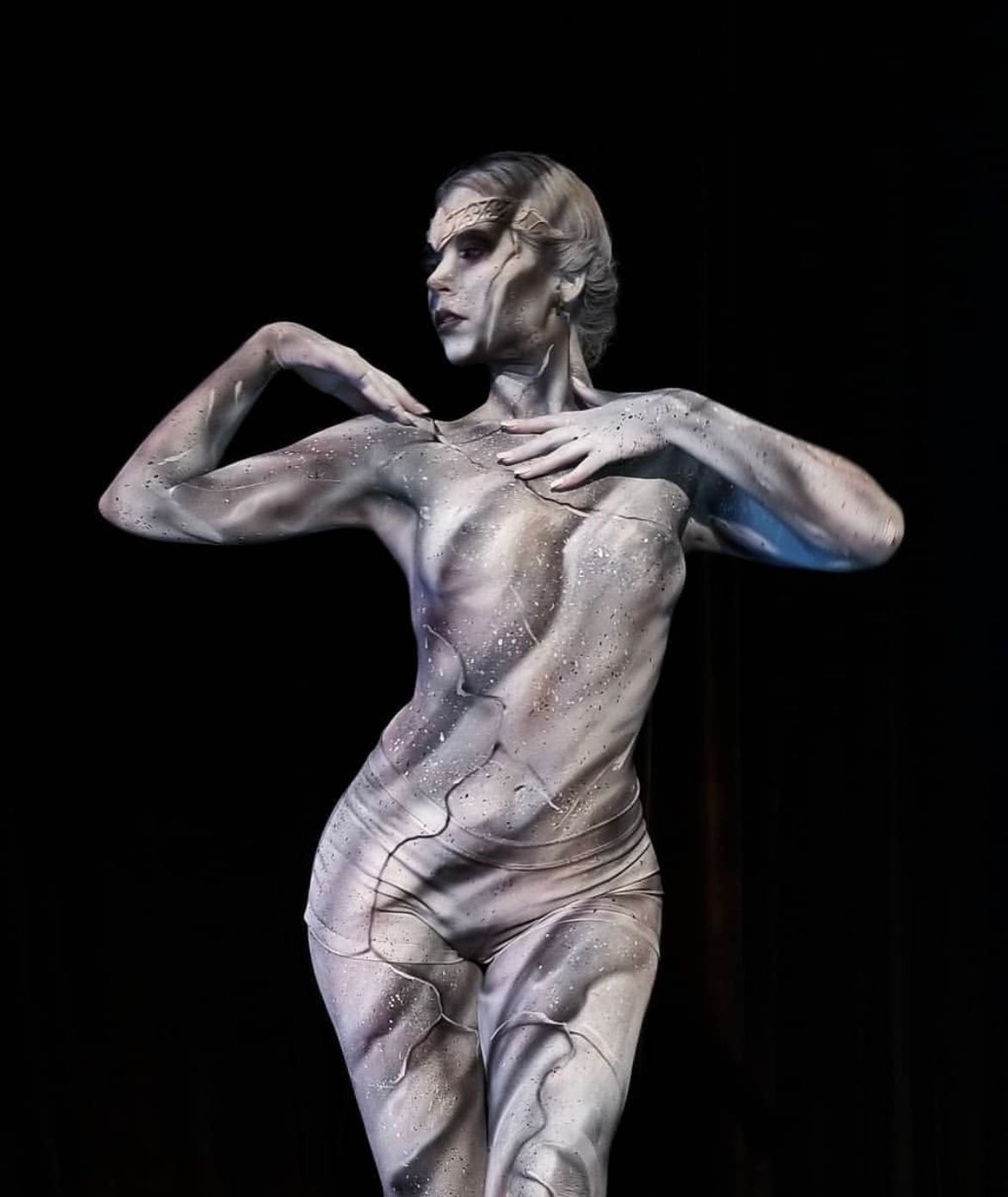 A woman with her body painted like a statue is standing in front of a black background.