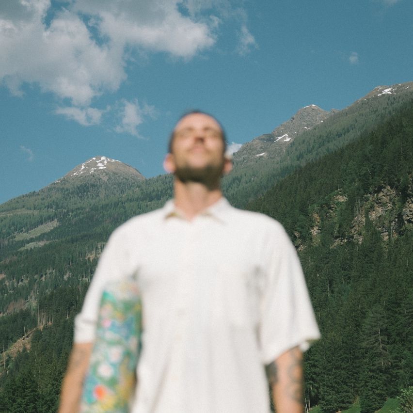 Blurred person in white shirt standing before forested mountains under a blue sky