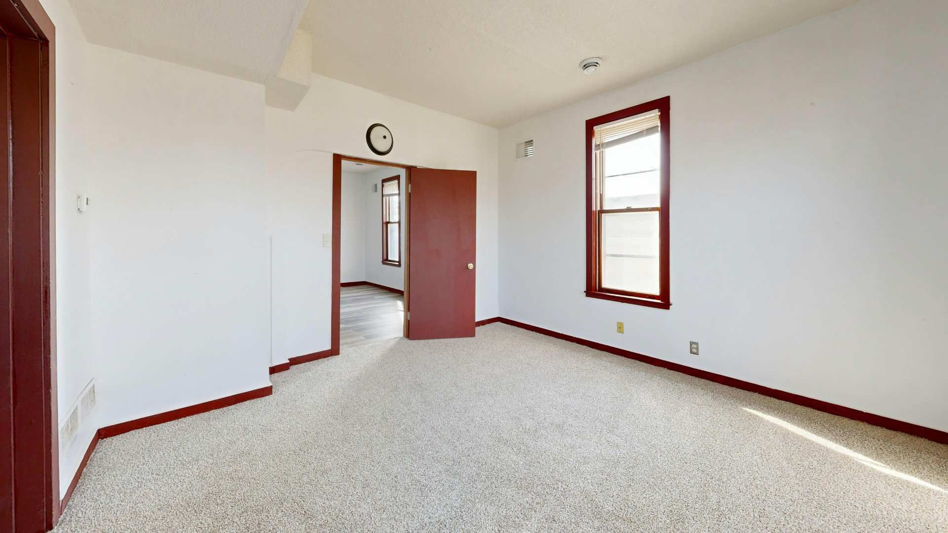 Empty beige bedroom with white walls, carpet, doorway, and a window with red trim