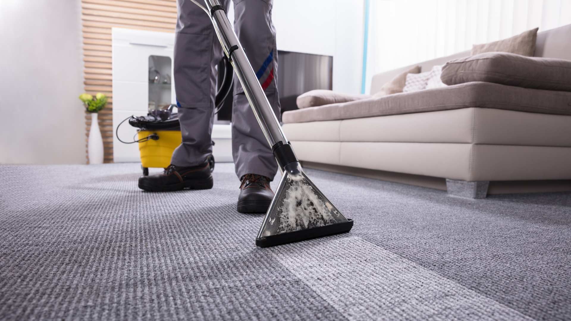 Person vacuuming a gray carpet in a bright living room with a yellow vacuum cleaner.