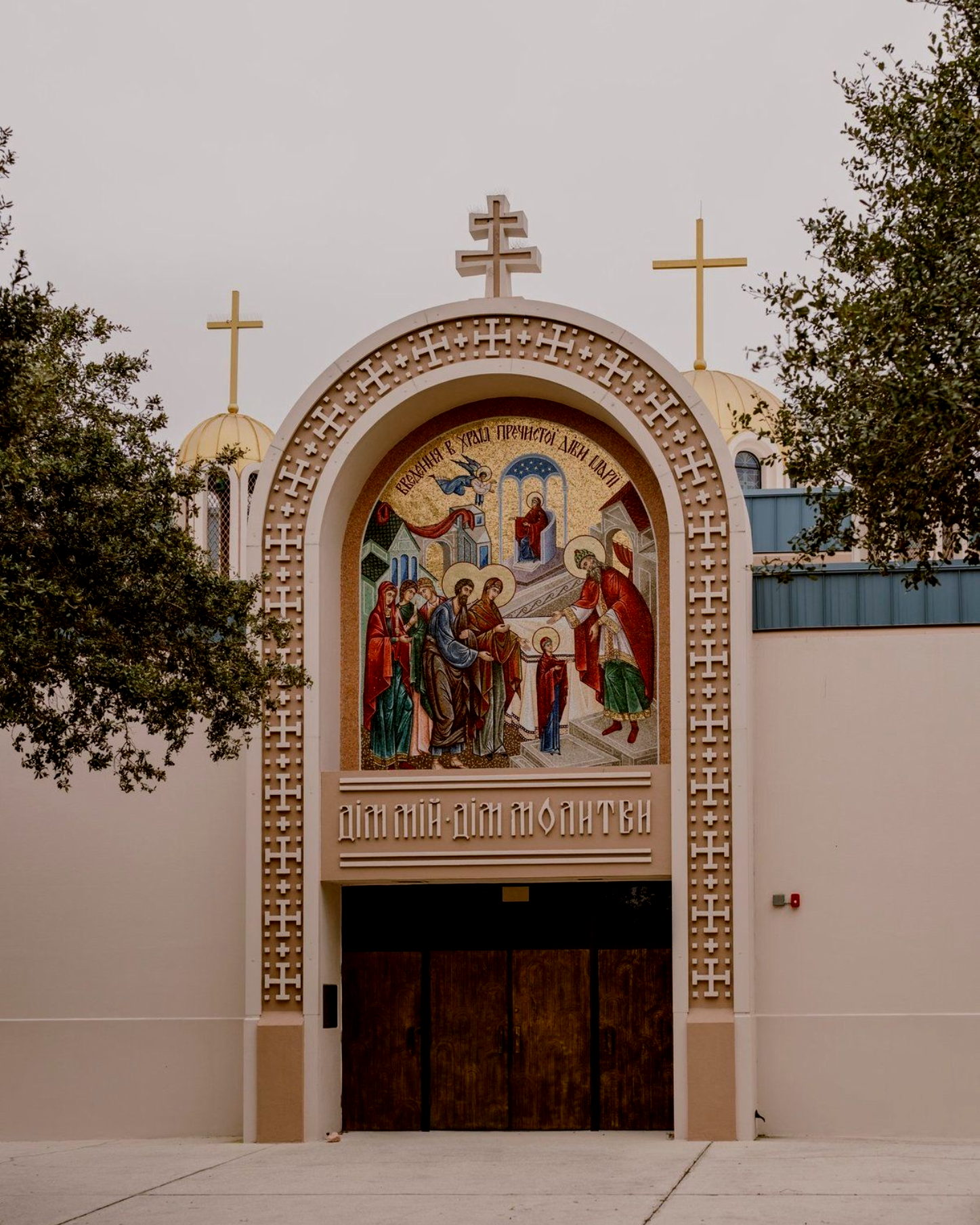 Church entrance with arched mosaic mural and crosses on top, framed by trees and a beige facade