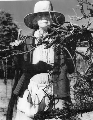 Woman wearing a hat and jacket, standing behind thorny branches, outdoors.