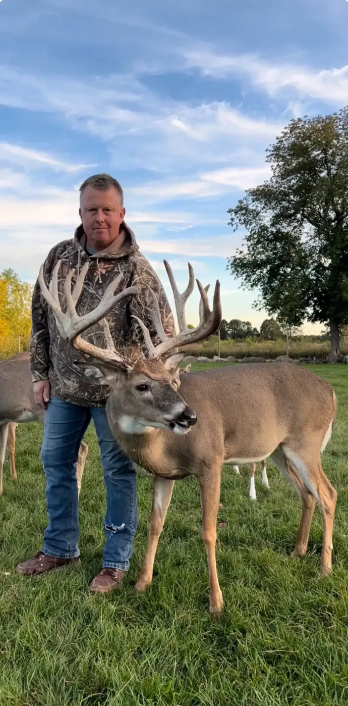 Man standing beside a deer with large antlers in a grassy field under a blue sky