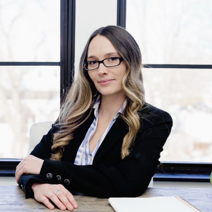 Woman in glasses seated at a desk by a window, wearing a black blazer and smiling softly.