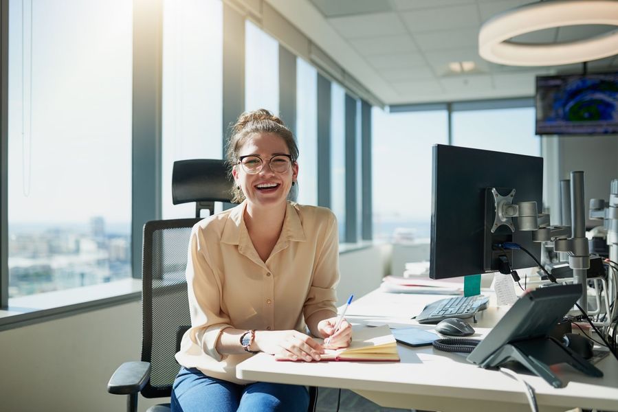 Smiling woman in beige blouse at a desk in a bright office with city views, laptop, papers, and phone.