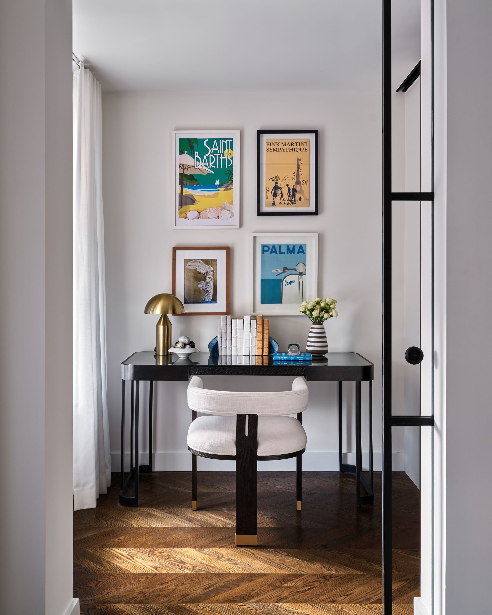 Compact home office with black desk, cream chair, four framed prints, and lamp in a bright hallway nook