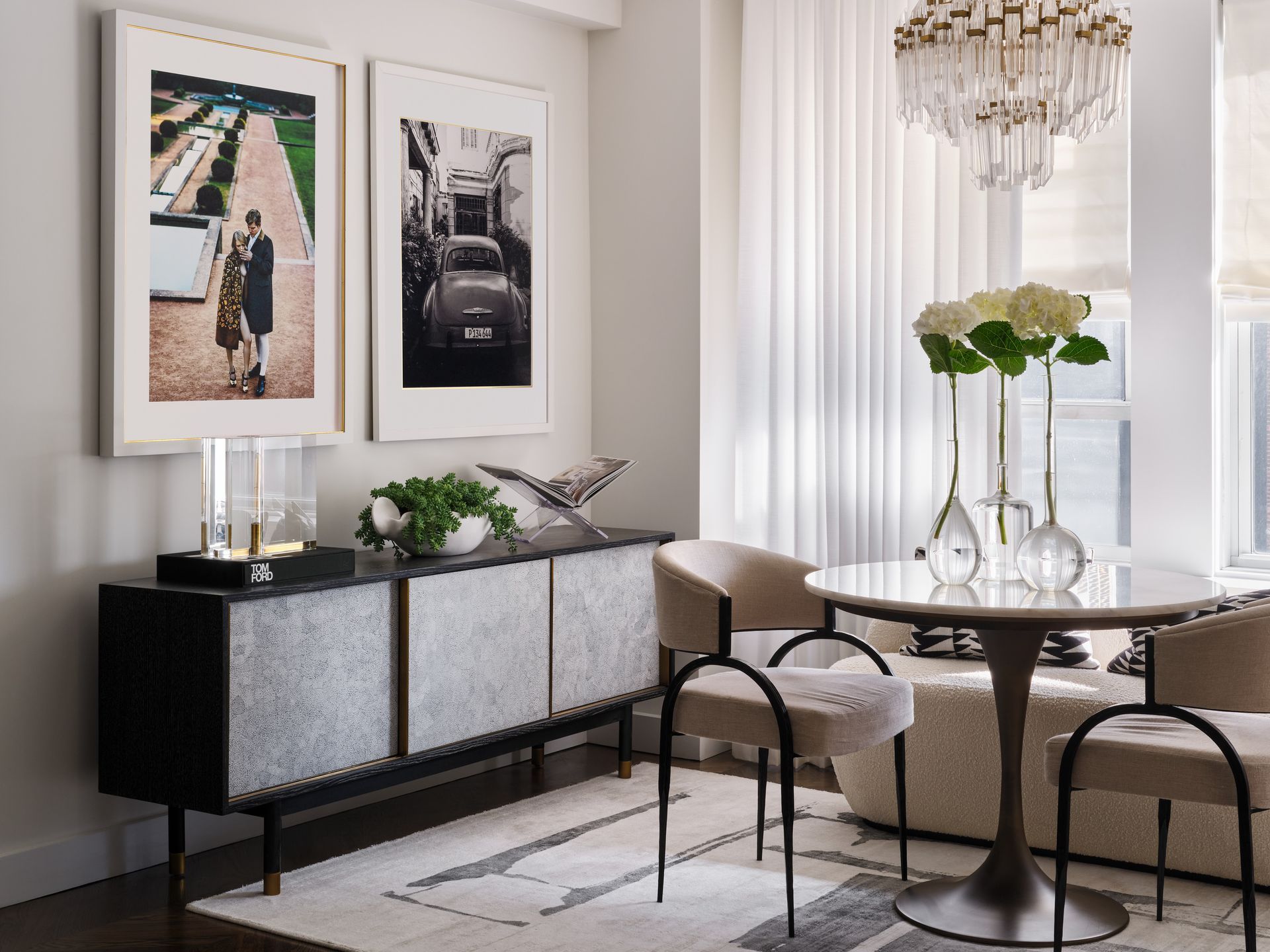 Elegant dining room with round glass table, upholstered chairs, black sideboard, and framed art.