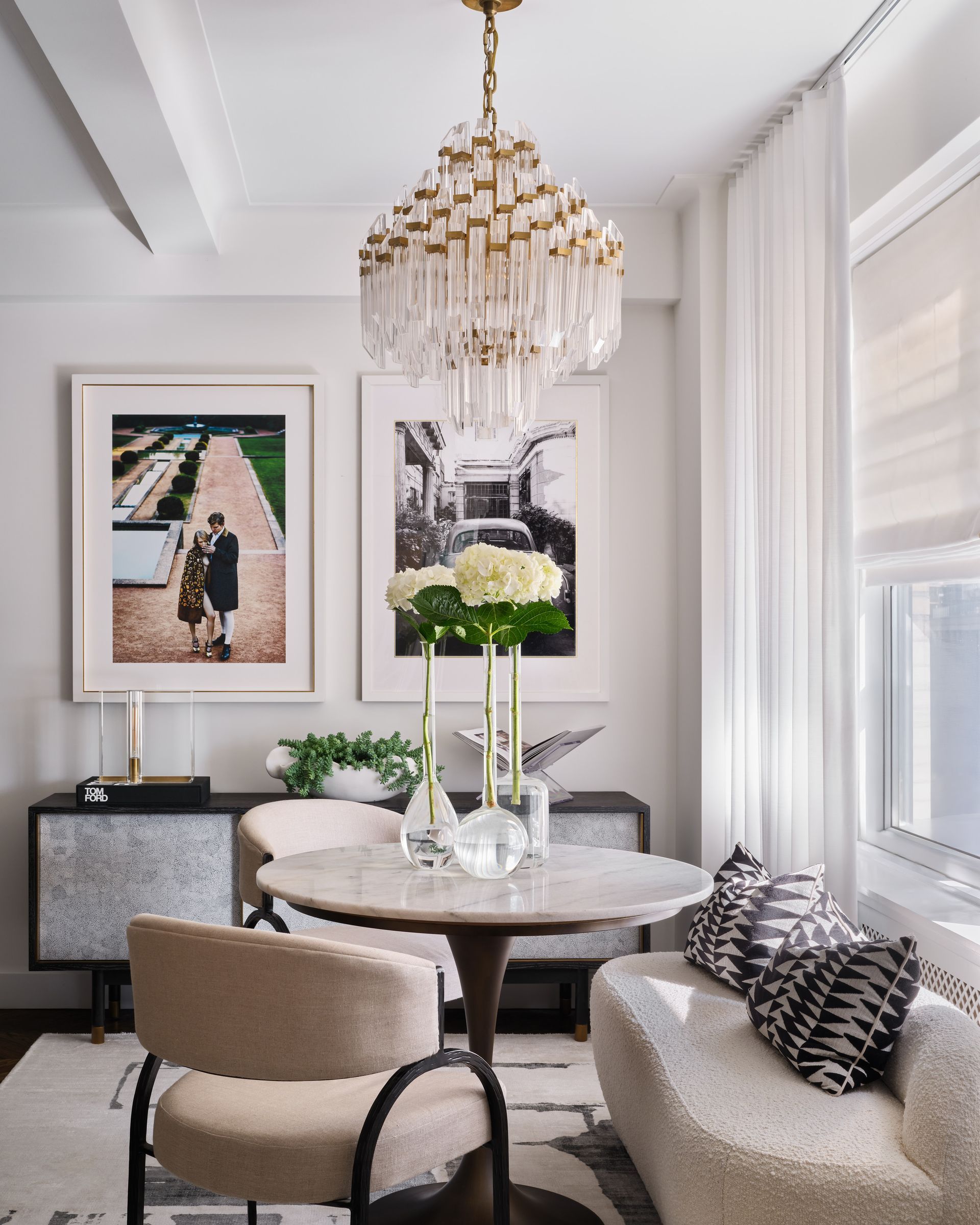Elegant dining nook with round table, beige chairs, chandelier, and framed art in a bright window-lined room