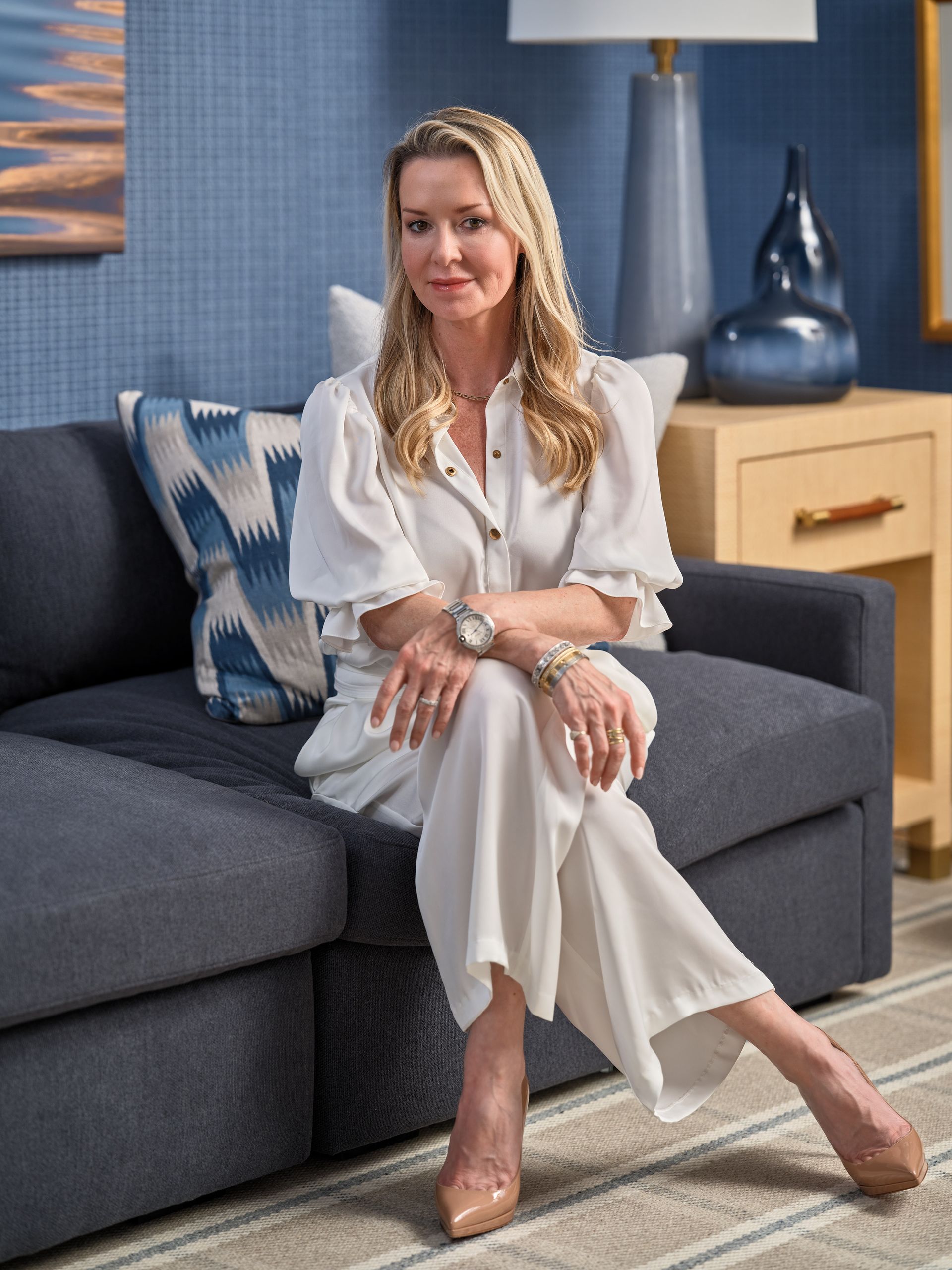 Woman seated on a gray sofa in a blue living room, wearing a white blouse and pants, looking at the camera.