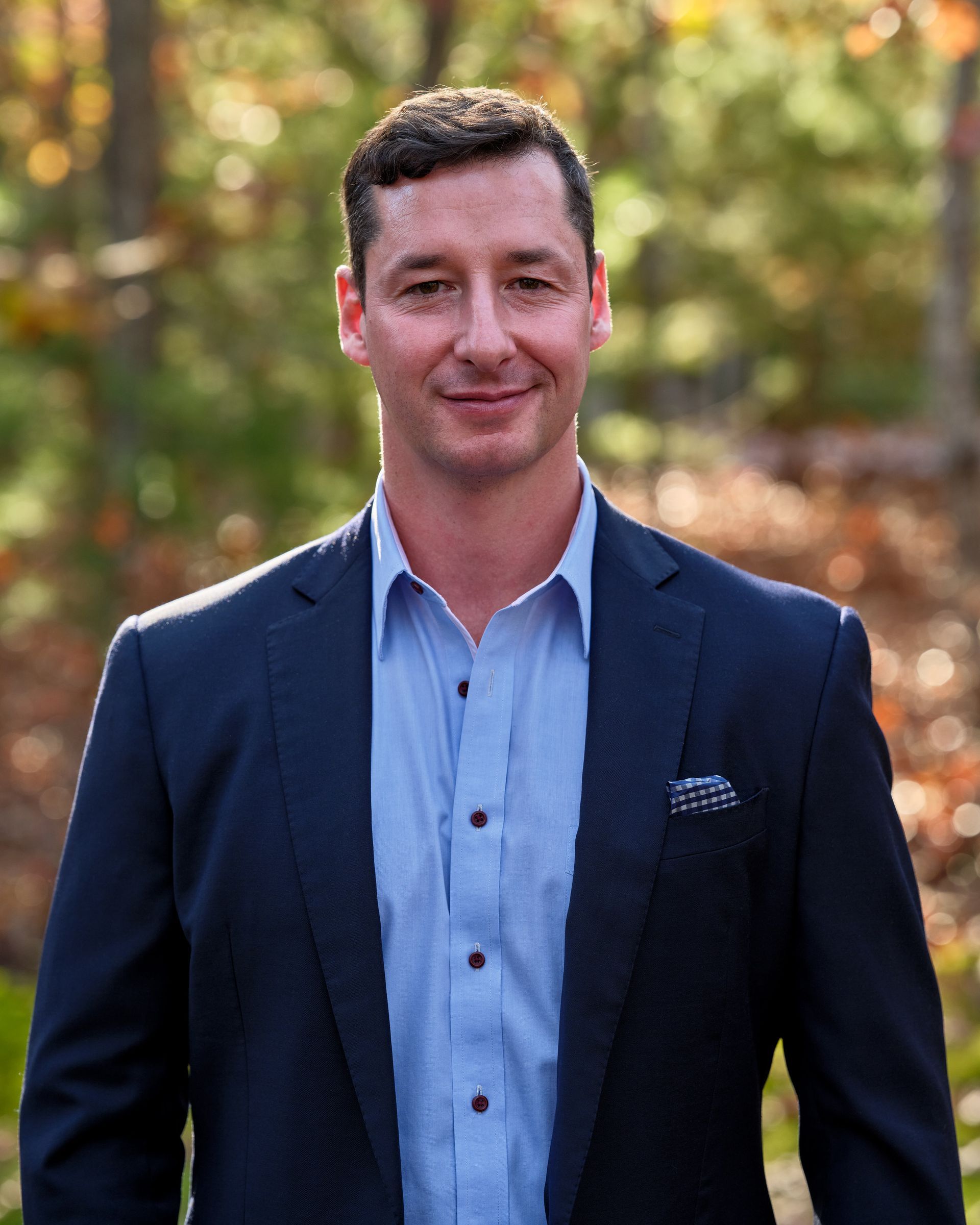 Man in a navy blazer and light blue shirt standing outdoors with a blurred autumn background