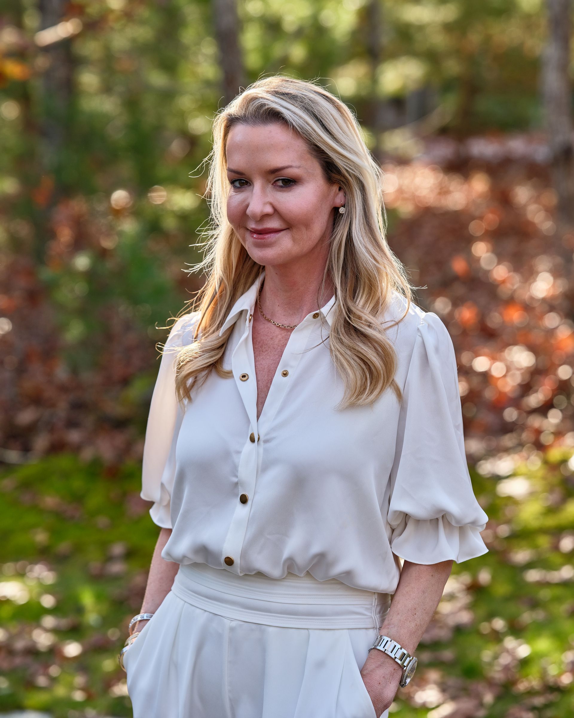 Woman in a white blouse and pants standing outdoors in a wooded setting with autumn foliage.