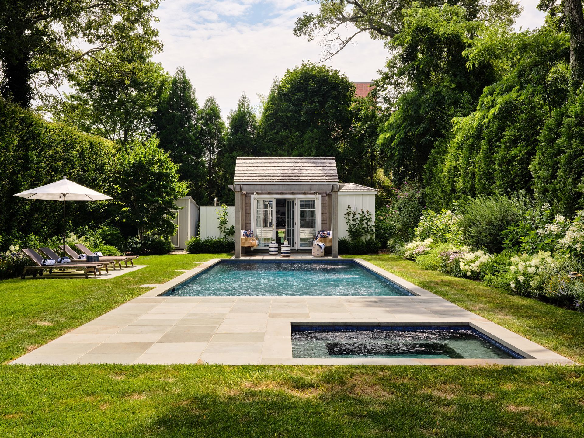 Modern backyard with a rectangular pool, lounge chairs, and a small cabana surrounded by lush greenery.