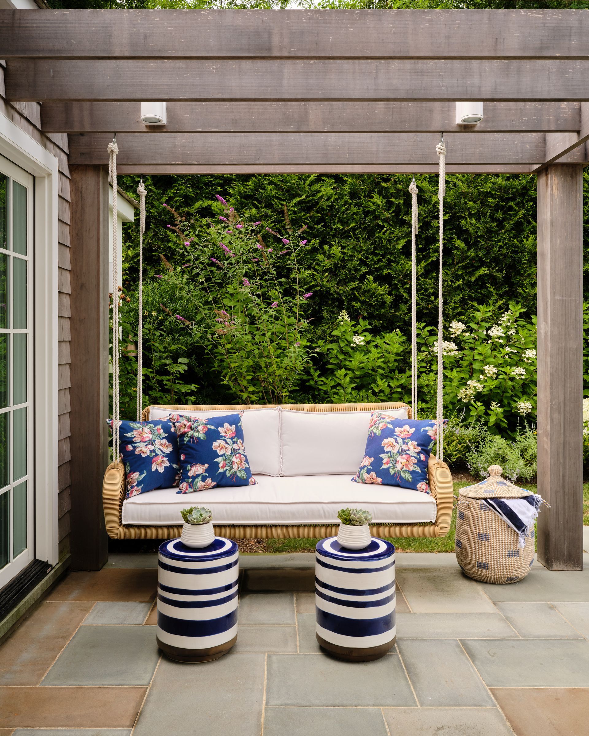 Patio with gray pergola, white cushioned sofa, blue floral pillows, and two striped ottomans