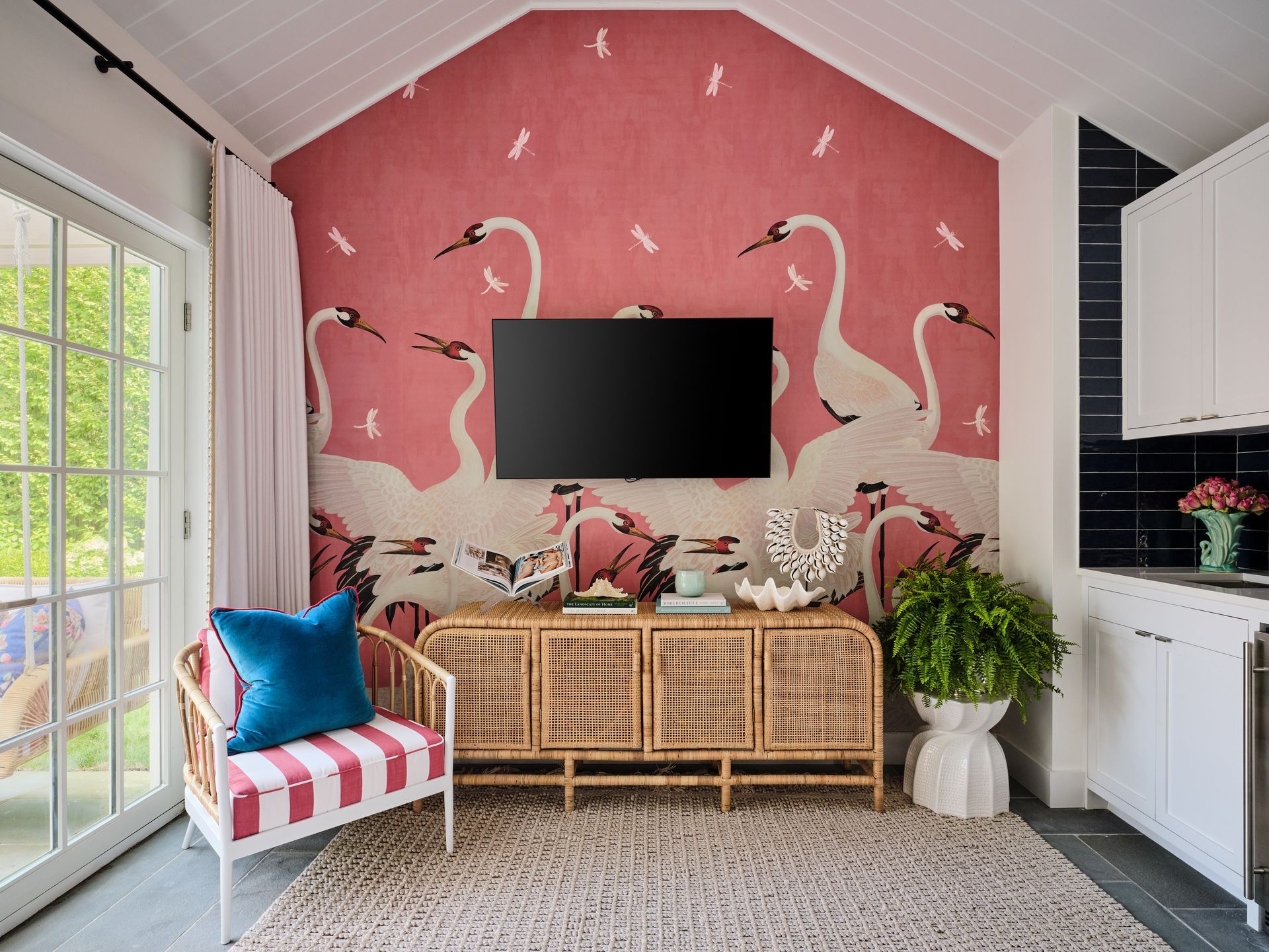 Bright pink sunroom with flamingo wallpaper, TV, wicker console, and colorful seating by large windows