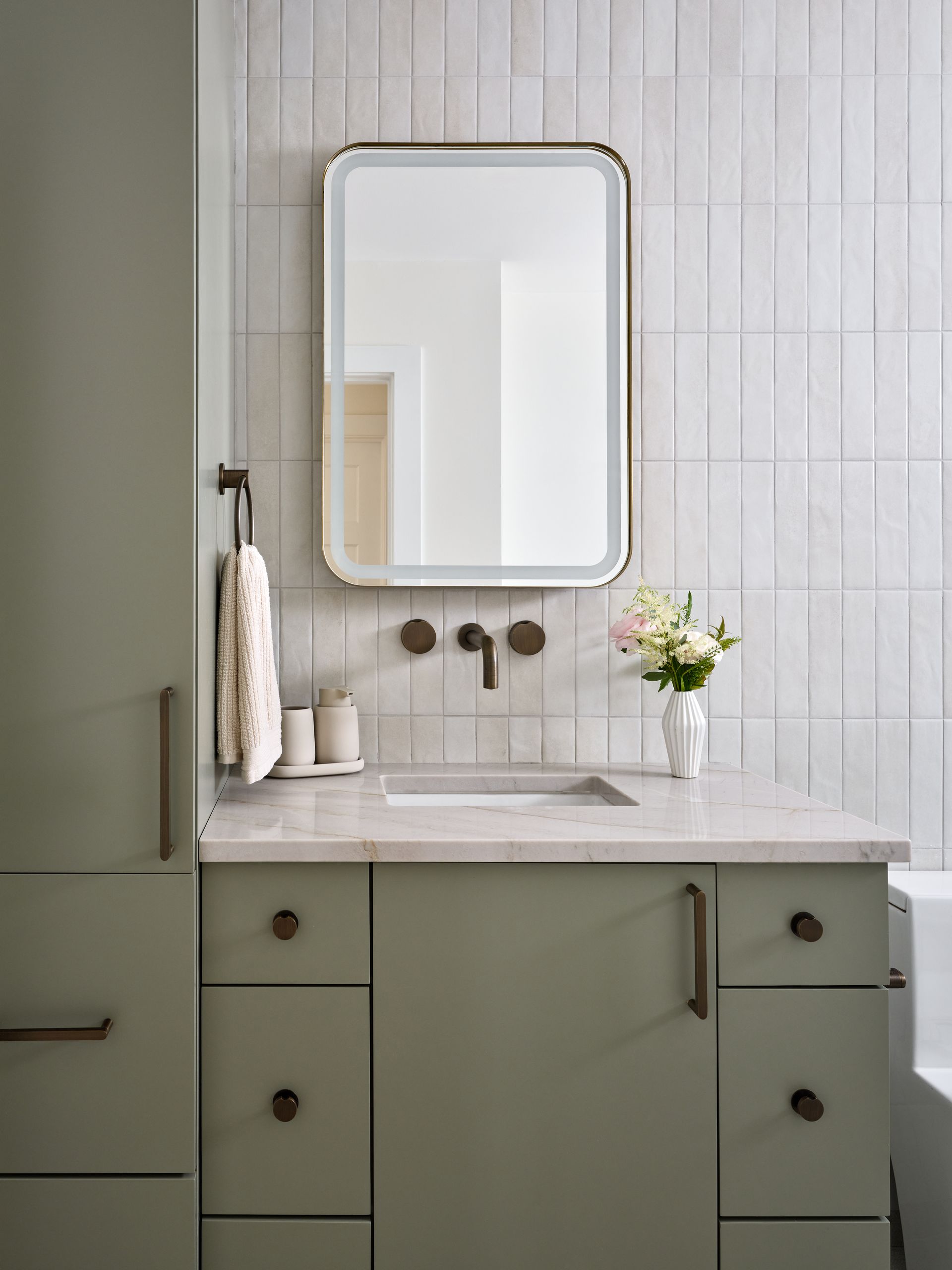 Modern bathroom vanity with green cabinets, marble countertop, round mirror, towel, and vase of flowers.