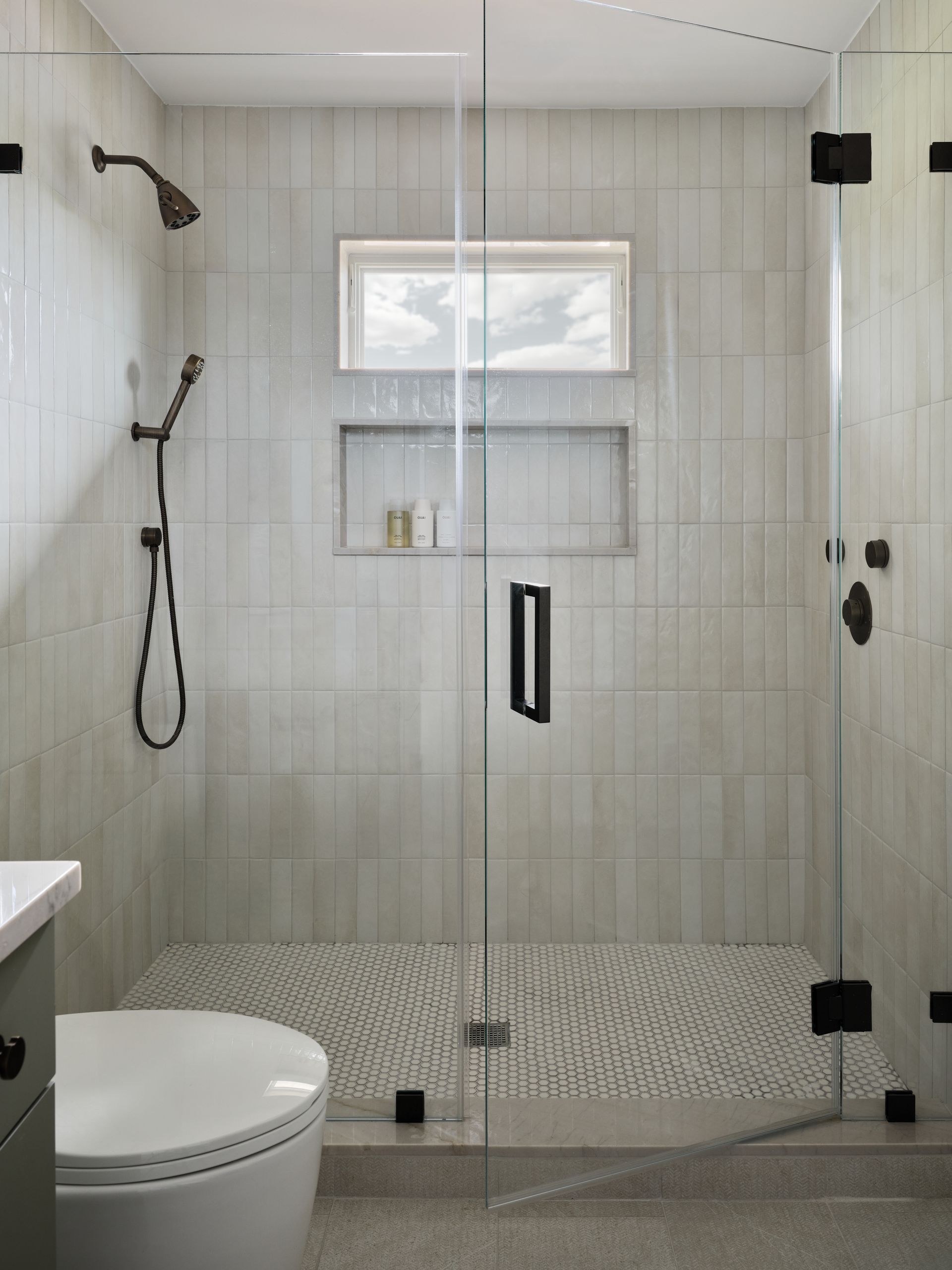 Modern bathroom shower with glass enclosure, white tile walls, bench, and toilet in the foreground