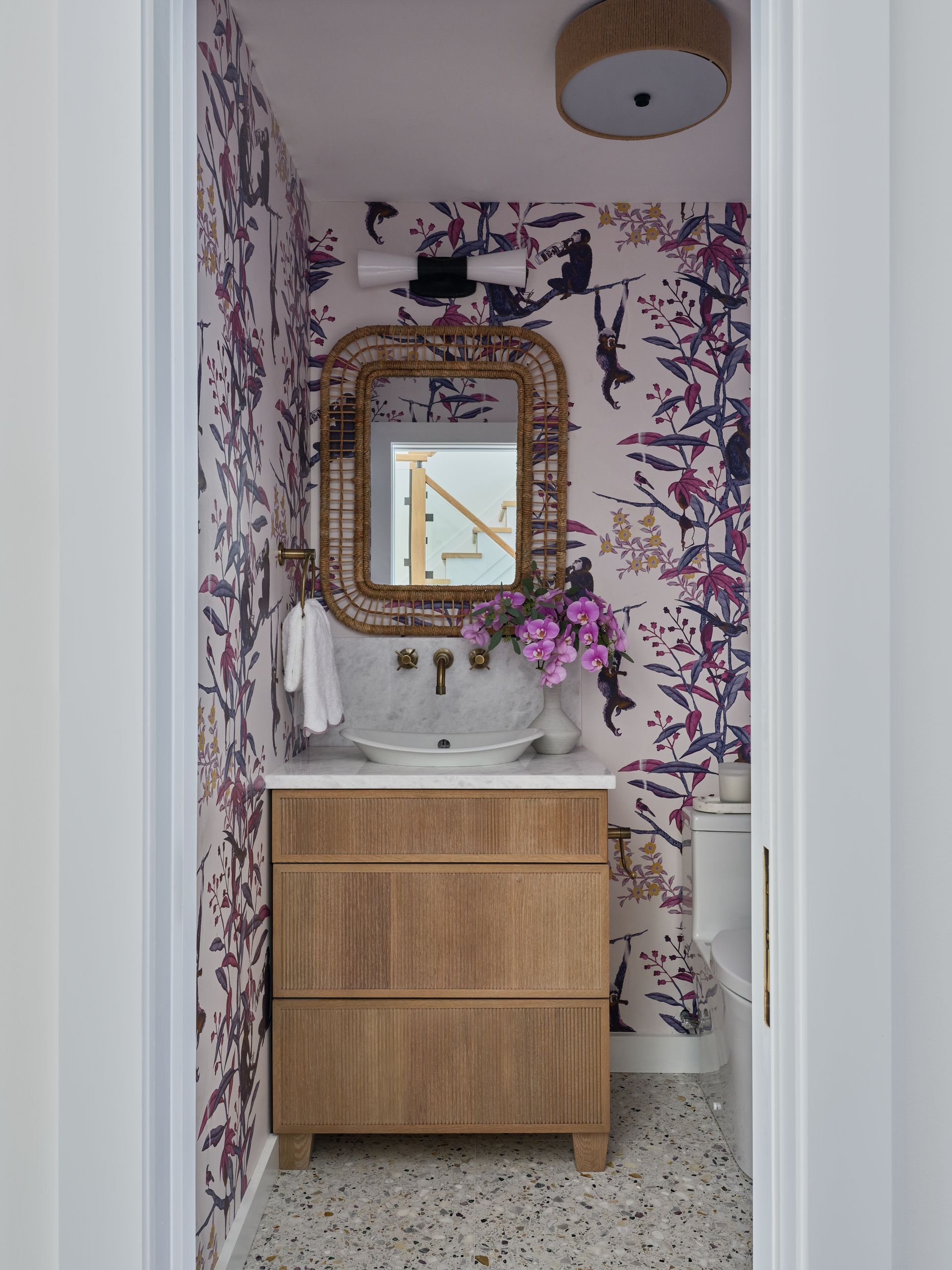 Small bathroom vanity with wood drawers, gold-framed mirror, purple floral wallpaper, and pink flowers.