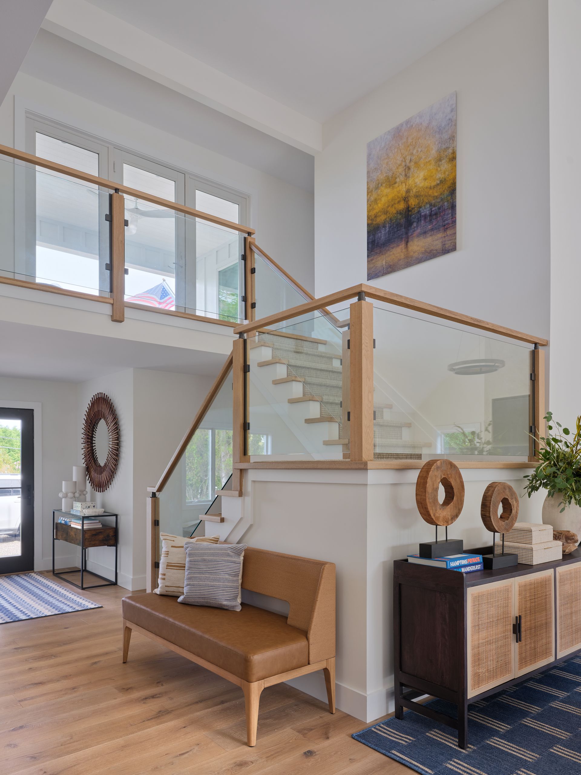Bright modern foyer with glass-railed staircase, wood bench, patterned rug, and framed wall art