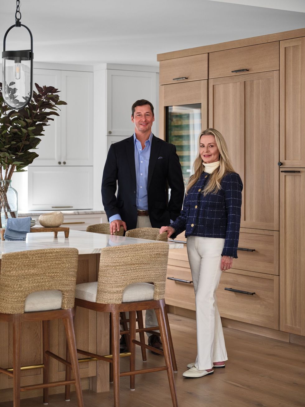 Two people standing in a bright kitchen with wood cabinets and a marble island.