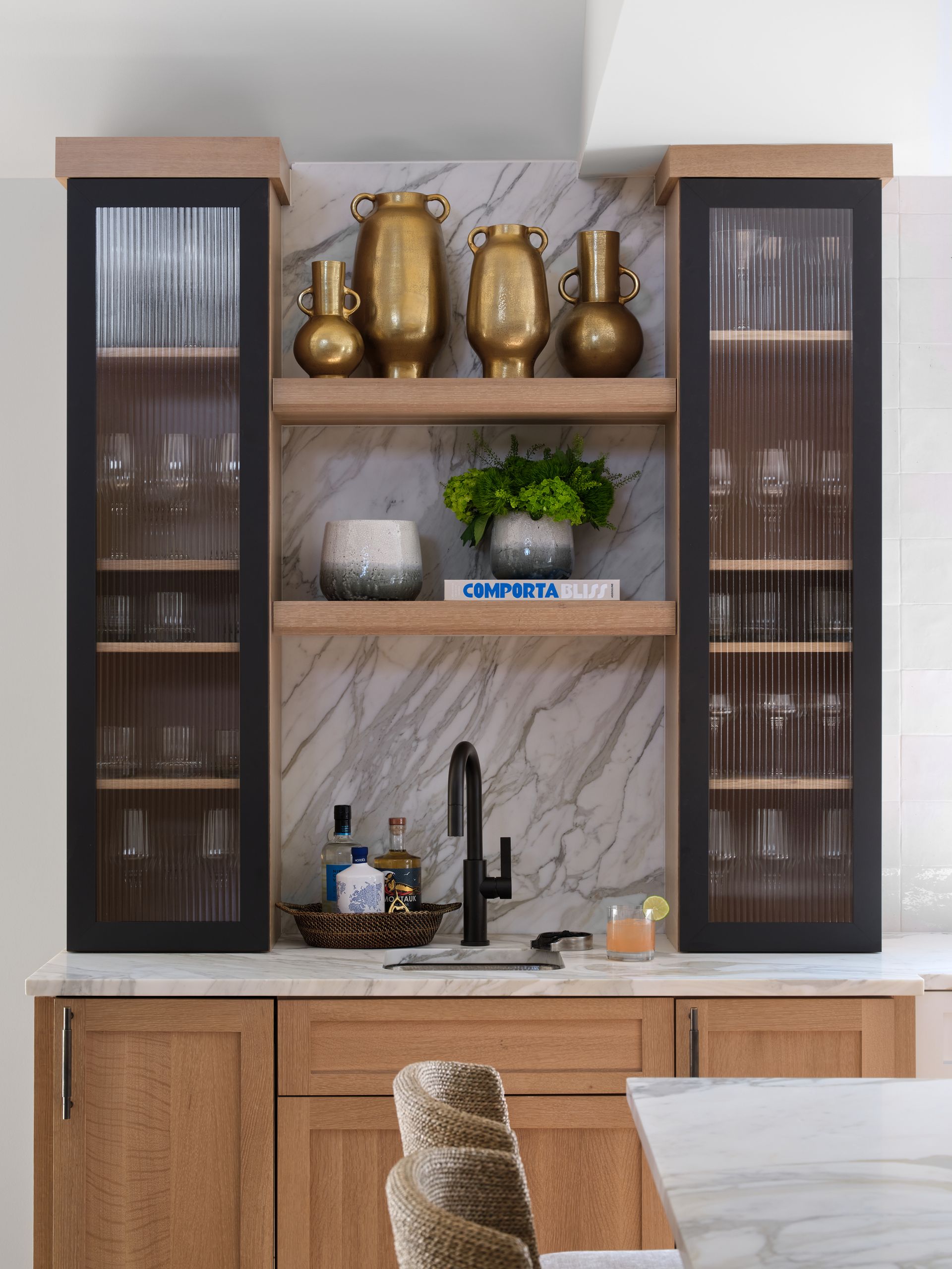 Modern built-in bar cabinet with wood doors, glass shelves, gold vases, and a sink beneath marble-look backing
