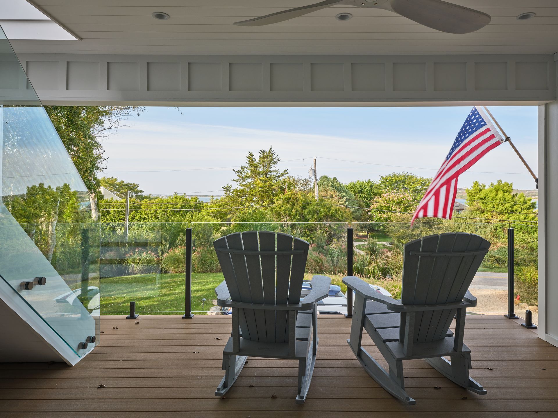 Two Adirondack chairs on a porch overlooking a green yard with an American flag.