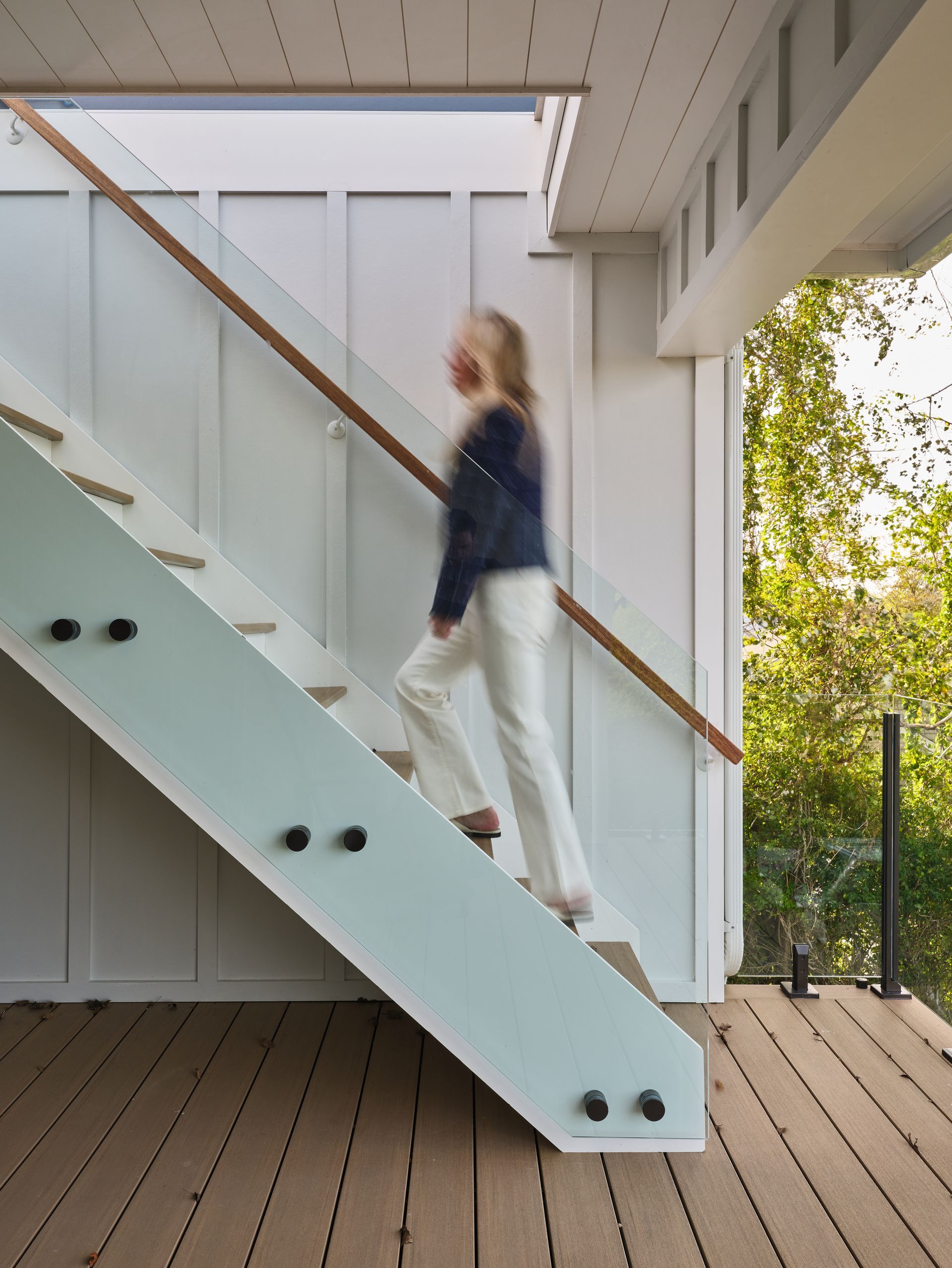 Person walking down white stairs on a wooden deck beside a modern house, with trees outside.