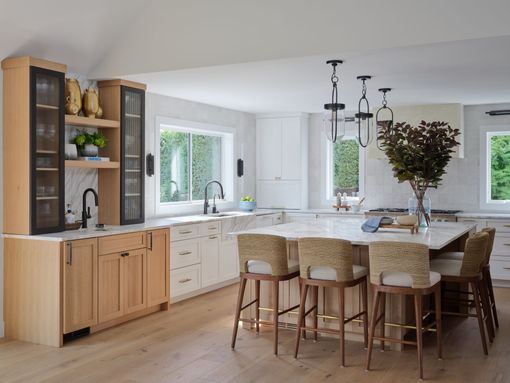 Bright modern kitchen with white island, wicker stools, wood cabinets, and large windows