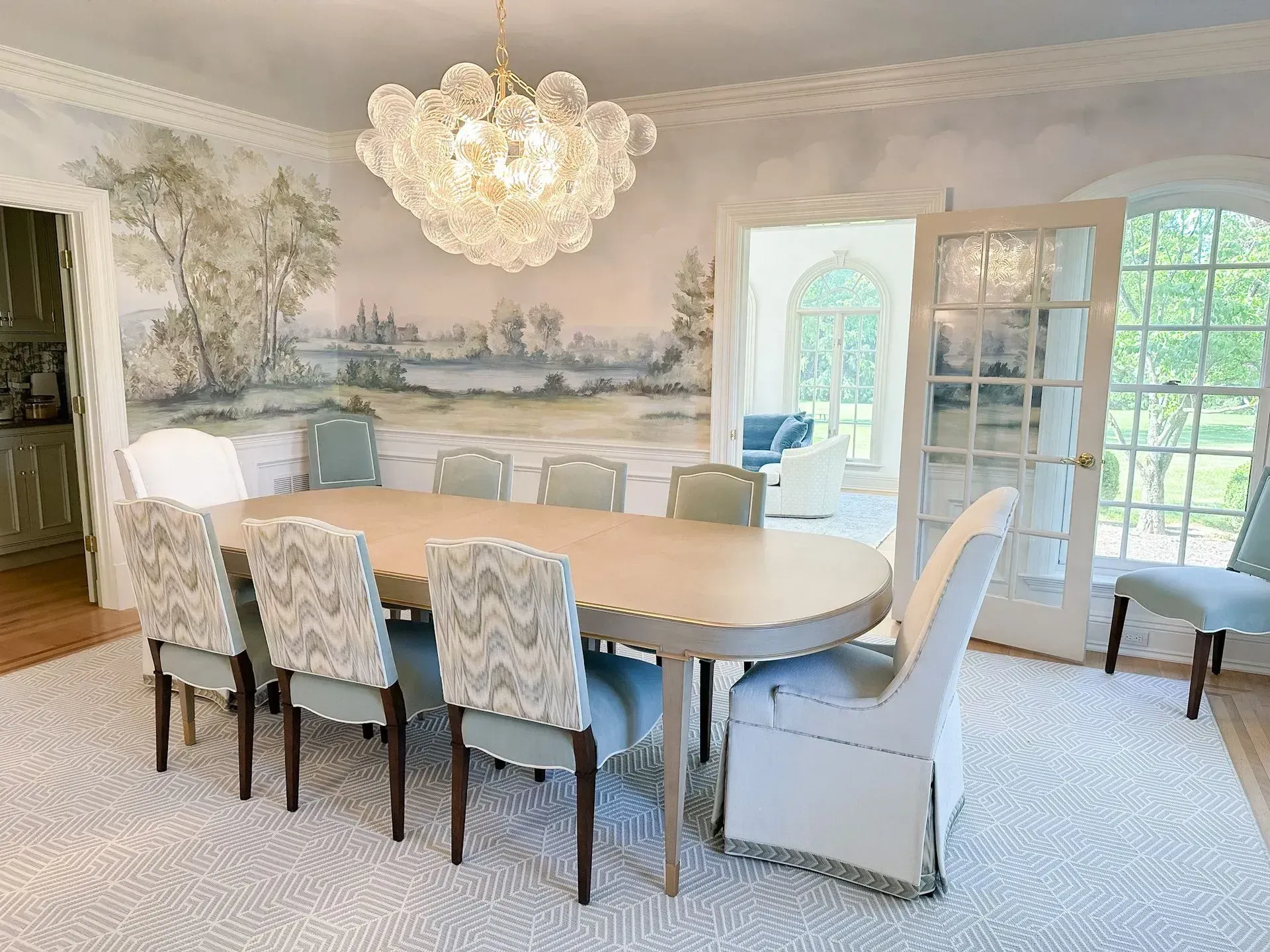 Elegant dining room with oval table, upholstered chairs, crystal chandelier, and scenic wall mural