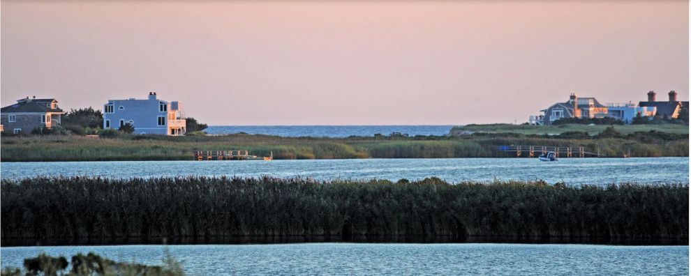 Coastal marsh at dusk with calm water, dark reeds, and distant white buildings under a pastel sky