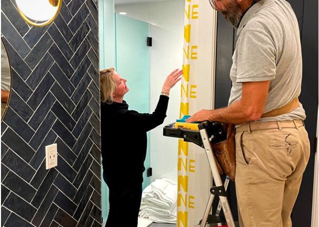 Two people install wall trim in a hallway, one on a ladder holding a tool.