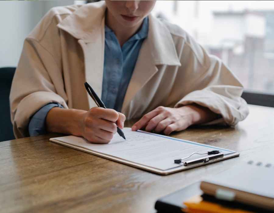 Person in a beige coat writing on a clipboard at a wooden table in a bright office setting