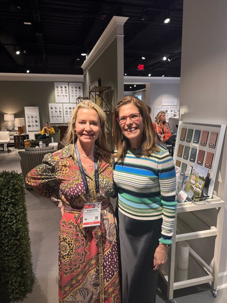 Two women smiling in an art gallery, standing beside a display wall and product samples.